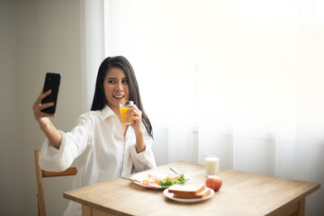 Obraz na płótnie Canvas Beautiful happy woman wearing a white shirt sitting and having a morning breakfast. plate of bread, an apple cup of milk. She holding an orange juice and taking a selfie by smartphone.