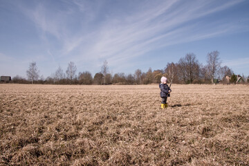 Naklejka na ściany i meble a little girl runs and plays in the field in the spring. a child in rubber boots plays with a stick