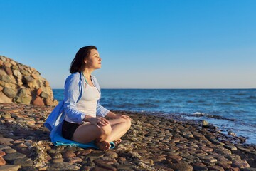 Fototapeta na wymiar Beautiful middle aged woman sitting in lotus position meditating on seashore at sunset