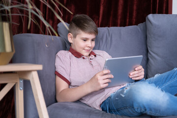 Young handsome teenager boy using tablet sitting on grey sofa at home