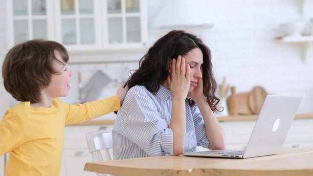 Nervous mom sits by table at home office during lockdown working on laptop trying to concentrate. Noisy child distracts from work, pulls the sleeve and hair, asking attention from annoyed busy mother 