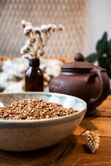 A plate with buckwheat groats on a wooden table. Clay teapot and dried flowers in the background. Healthy eating.