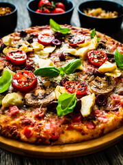 Pizza capricciosa with white mushrooms, ham, artichoke, tomatoes, olives, parmesan and mozzarella on wooden background
