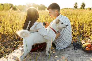 Stylish couple with white dog relaxing on blanket in sunny light in summer meadow. Picnic with pet