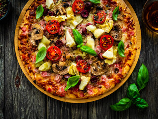 Pizza capricciosa with white mushrooms, ham, artichoke, tomatoes, olives, parmesan and mozzarella on wooden background
