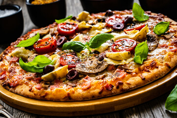 Pizza capricciosa with white mushrooms, ham, artichoke, tomatoes, olives, parmesan and mozzarella on wooden background
