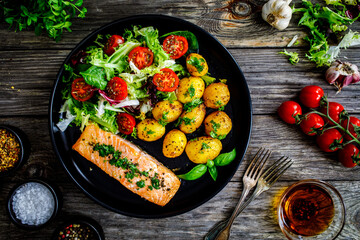Roasted salmon steak with roast potatoes and vegetable salad served on black plate on wooden table

