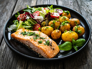 Roasted salmon steak with roast potatoes and vegetable salad served on black plate on wooden table
