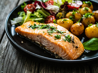 Roasted salmon steak with roast potatoes and vegetable salad served on black plate on wooden table
