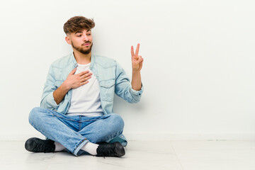 Young Moroccan man sitting on the floor isolated on white background taking an oath, putting hand on chest.