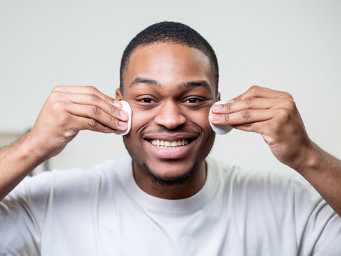 Male facial care. Skin cleansing. Hygiene procedure. Cheerful fun African man in white t-shirt using cotton pad with tonic smiling on light background.