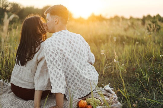 Stylish couple kissing on blanket in sunny light among grass in summer meadow, enjoying sunset
