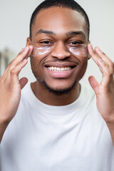 Male skin care. Facial treatment. Metrosexual lifestyle. Happy cheerful African man in white t-shirt enjoying applying hydrogel face eye patches smiling.