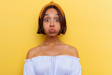 Young mixed race woman isolated on yellow background blows cheeks, has tired expression. Facial...
