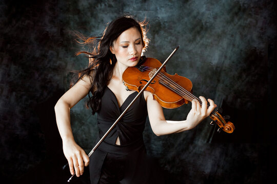 An Asian young woman is playing violin. Violinist is performing on the stage.  Her hair  flying in contour light. Emotional. Musical Instrument
