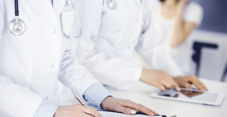 Unknown woman-doctors at work with patient at the background. Female physicians filling up medical documents or prescription while standing in hospital reception desk, close-up. Health care concept