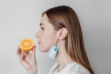 Loss of smell concept. Caucasian young woman in a medical mask sniffing an orange on a white...