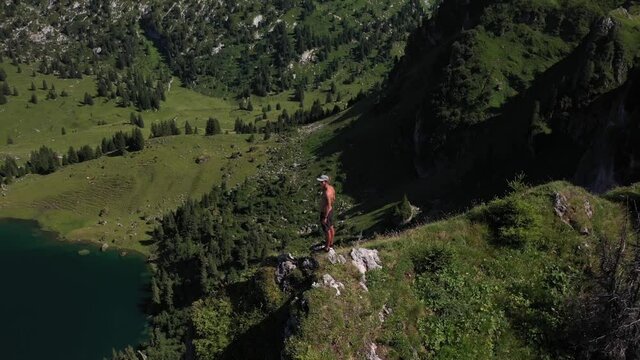A young, fit and strong man is standind alone by himself on the edge of a cliff, controlling his drone and looks up to enjoy the view during the hot sunny day in Seebergsee, Switzerland.