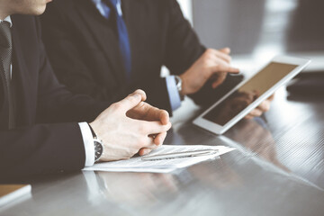 Unknown businessmen using tablet computer and work together at the glass desk in modern office, close-up. Teamwork and partnership concept