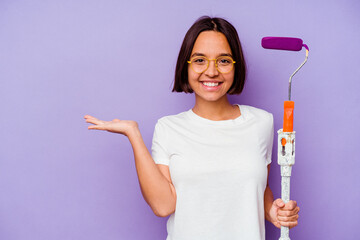 Young painter mixed race woman holding a paint stick isolated on purple background showing a copy space on a palm and holding another hand on waist.