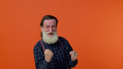 Mature old bearded gray-haired grandfather in shirt shouting, raising fists in gesture I did it, celebrating success, winning and goal achievement. Senior man on orange background. People lifestyle