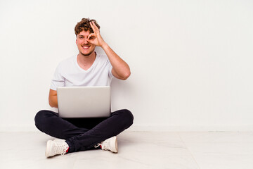 Young caucasian man sitting on the floor holding on laptop isolated on white background excited keeping ok gesture on eye.