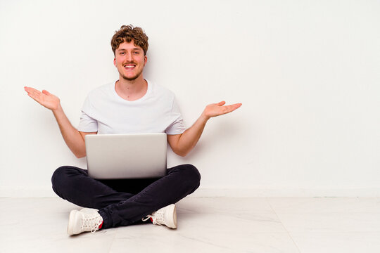 Young caucasian man sitting on the floor holding on laptop isolated on white background makes scale with arms, feels happy and confident.