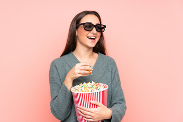 Young caucasian woman isolated on pink background with 3d glasses and holding a big bucket of popcorns