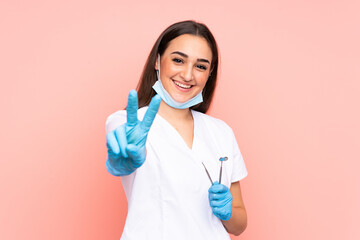 Woman dentist holding tools isolated on pink background smiling and showing victory sign