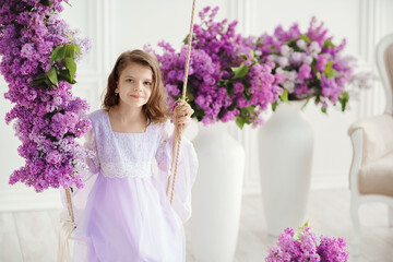 Beautiful little girl of preschool age in a delicate dress sitting on a swing decorated with flowers of lilac.