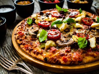 Pizza capricciosa with white mushrooms, ham, artichoke, tomatoes, olives, parmesan and mozzarella on wooden background
