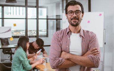 Closeup on caucasian beard handsome creative and business man wearing casual shirt and eyeglasses, crossing arms and smiling with blur background of colleagues working in indoor office or workplace.