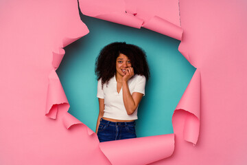 Young African American woman in torn paper isolated on blue background biting fingernails, nervous and very anxious.