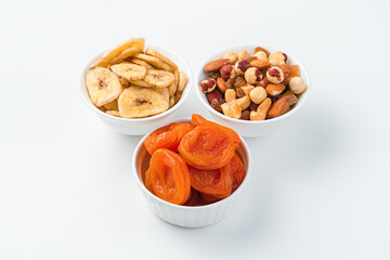 Dried apricots, banana chips and a mixture of nuts in three bowls on a white background.