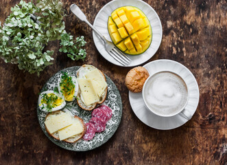 Delicious brunch, breakfast - cappuccino, fresh mango, boiled egg with micro greens, salami, cheese sandwiches on a wooden background, top view