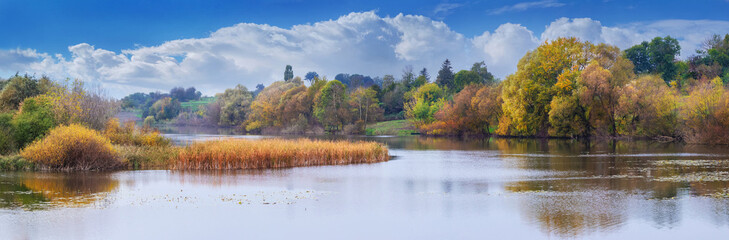 Autumn landscape with colorful trees by the river and picturesque blue sky with white clouds, panorama