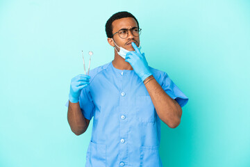 African American dentist holding tools over isolated blue background having doubts while looking up
