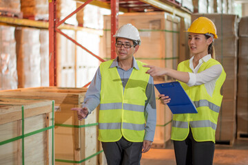 Young Asian woman worker use clip board and man engineer supervisor walking to check product box shelf in big warehouse factory store which smile and felling happy, logistic and transportation concept