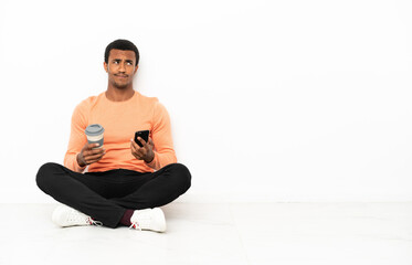 African American man sitting on the floor over isolated copyspace background holding coffee to take away and a mobile while thinking something