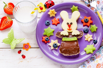 Fun food for kids - cute smiling bunny in a magic hat for a healthy breakfast with a chocolate spread sandwich and fresh banana, apple, kiwi, strawberry and blueberries served with a glass of milk