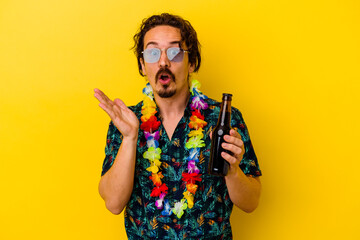 Young caucasian man wearing a hawaiian necklace holding a beer isolated on yellow background surprised and shocked.