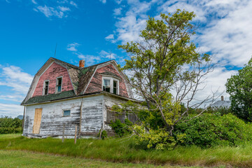 Fototapeta na wymiar Old, abandoned pink house on the prairies with trees, grass, and blue sky in Verwood, Saskatchewan, Canada