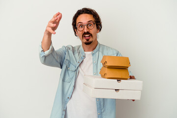 Young caucasian man holding hamburger an pizzas isolated on white background receiving a pleasant surprise, excited and raising hands.
