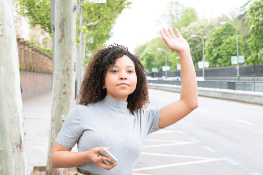 Woman With Curly Hair Hailing A Cab Or Uber With Raised Hand While Holding A Cell Phone In The City. Horizontal Photograph.