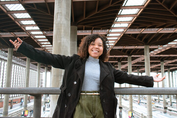 Happy curly-haired woman smiling and spreading arms with a train station behind.