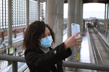 Portrait of young woman with sirurgical mask taking selfie with smartphone on train station of background.