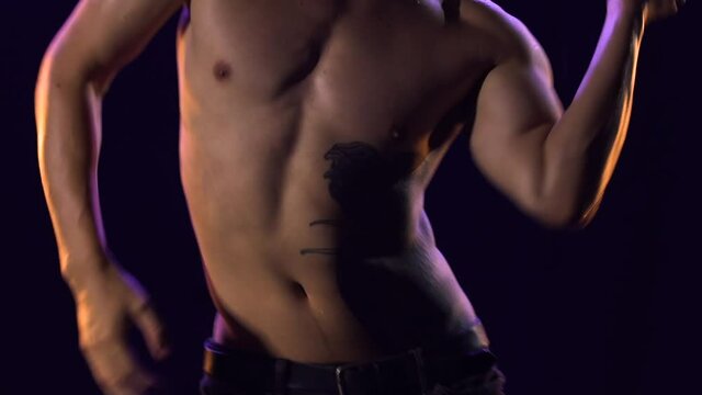 Handsome young man dancing elements of modern ballet, splitting raindrops. Male dancer with naked torso moves plastically on surface of the water in dark studio among splashes. Close up. Slow motion.