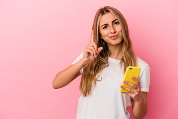 Young blonde caucasian woman holding a yellow mobile phone isolated showing number one with finger.