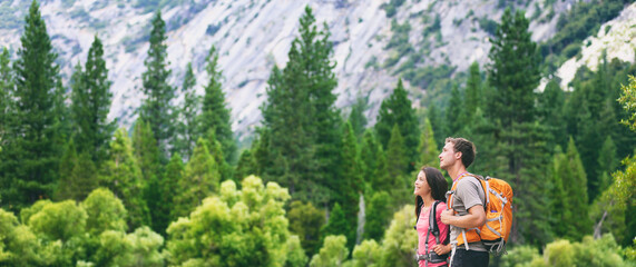 Hikers hiking in outdoor mountain nature landscape looking away to the side copy space on green pine trees forest. Backpacking tourists woman and man couple with backpacks walking outside banner.