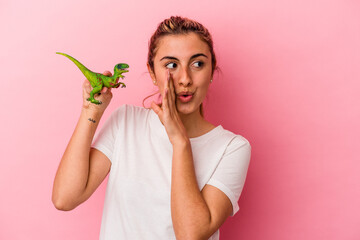 Young blonde caucasian woman holding a dinosaur miniature isolated on pink background is saying a...
